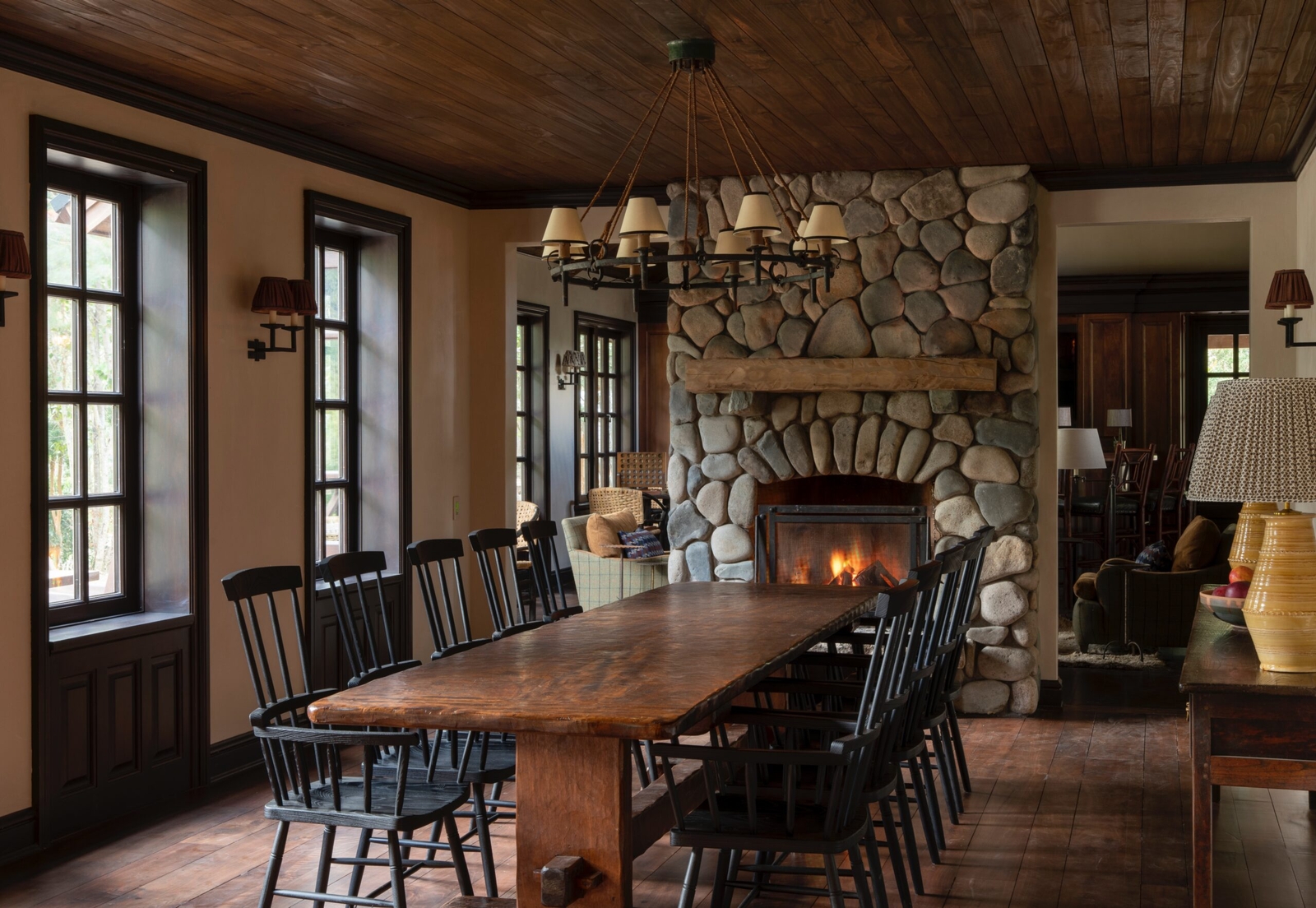 Cozy dining room with a large wooden table, black chairs, and a stone fireplace lit with fire.