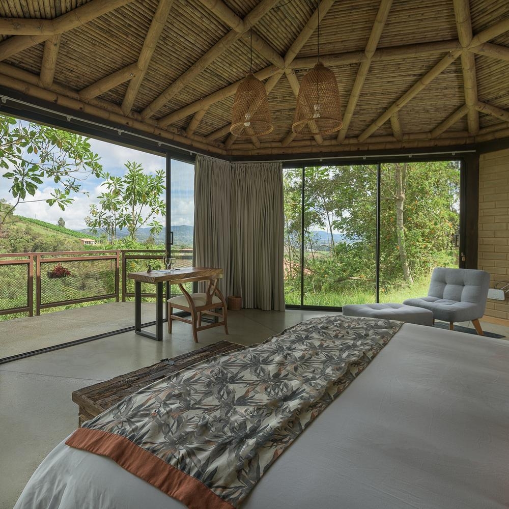 Rustic bedroom with thatched roof, large window overlooking greenery, and modern furniture.