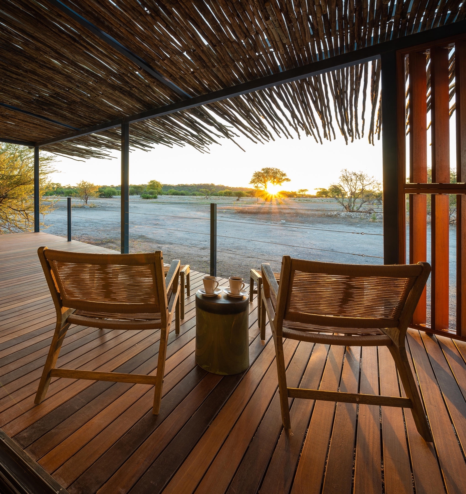 A serene wooden patio with two chairs and a small table overlooking a sunset in a peaceful landscape.