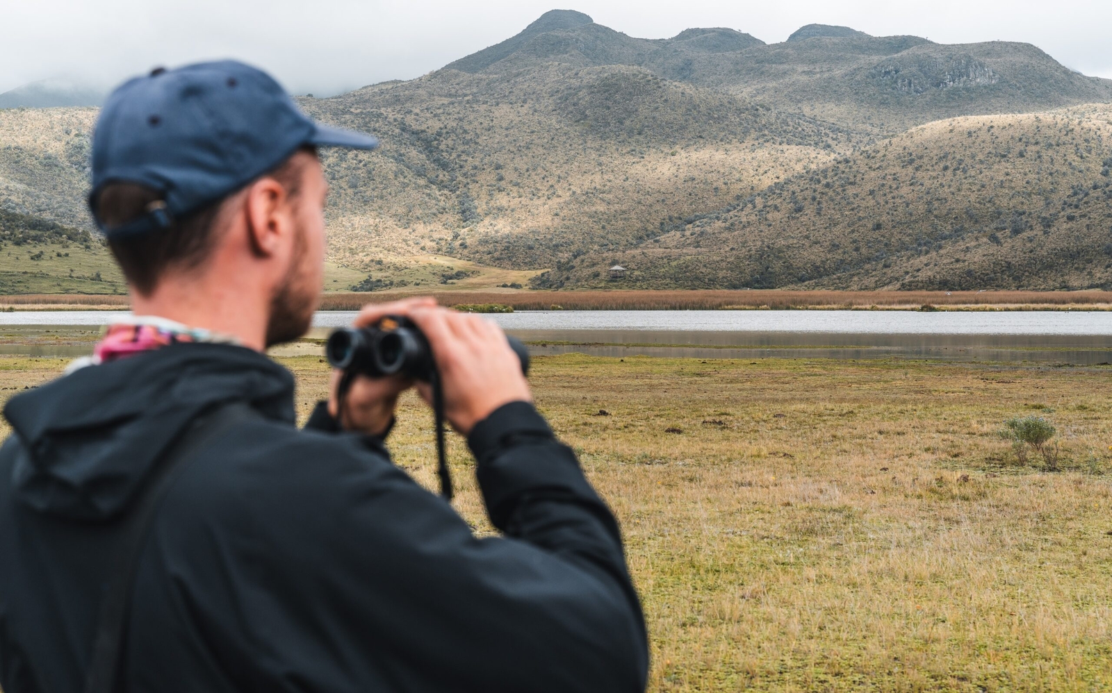 Man with binoculars looking at a mountainous landscape across a lake.