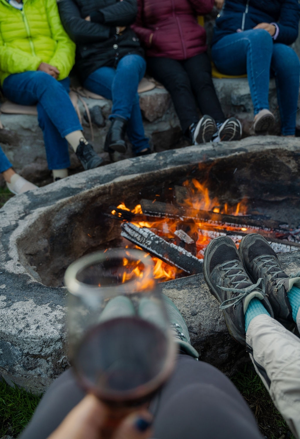 Person with a wine glass foreground, feet propped near a campfire with others in background.