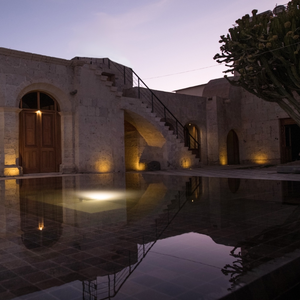Illuminated stone building with arches and a reflection on a pool at dusk.