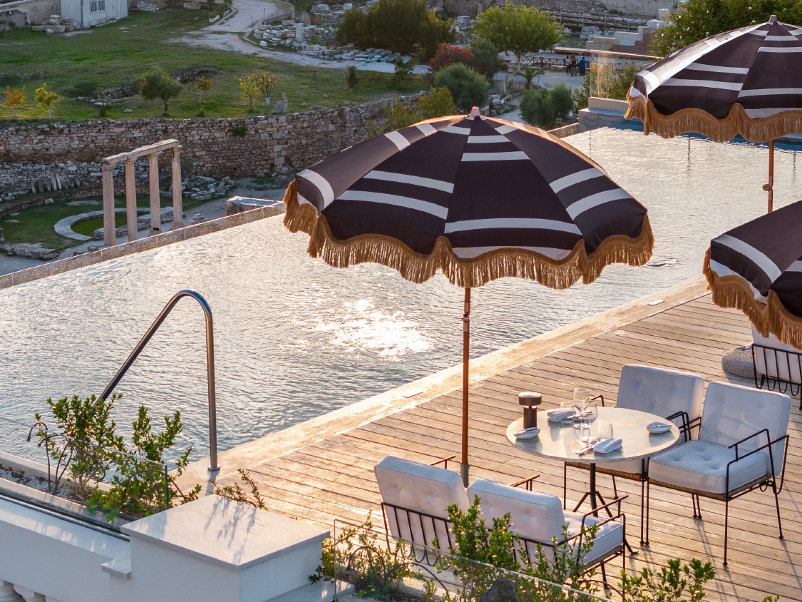 A luxury poolside scene featuring a tranquil water surface, elegant dining table, and shaded umbrellas overlooking lush greenery and ruins.