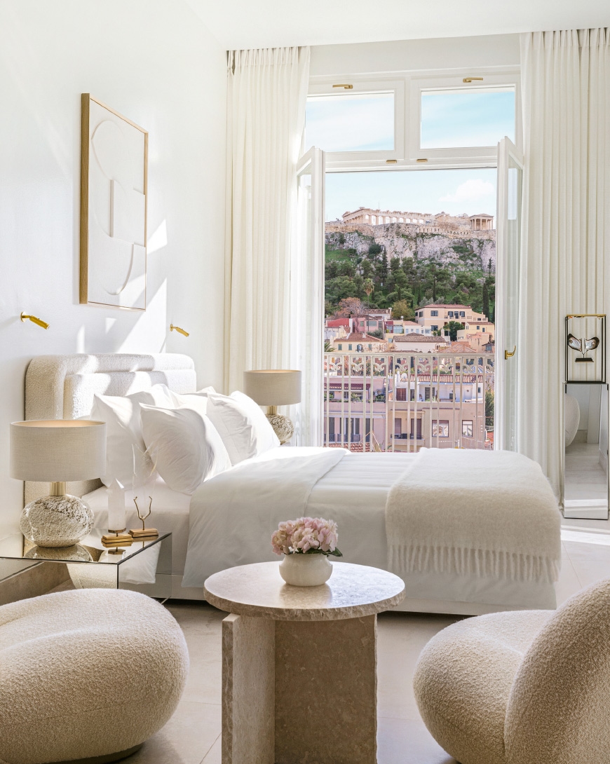 Bright bedroom featuring modern decor, soft textures, and a view of Athens with the Acropolis in the background through open windows.
