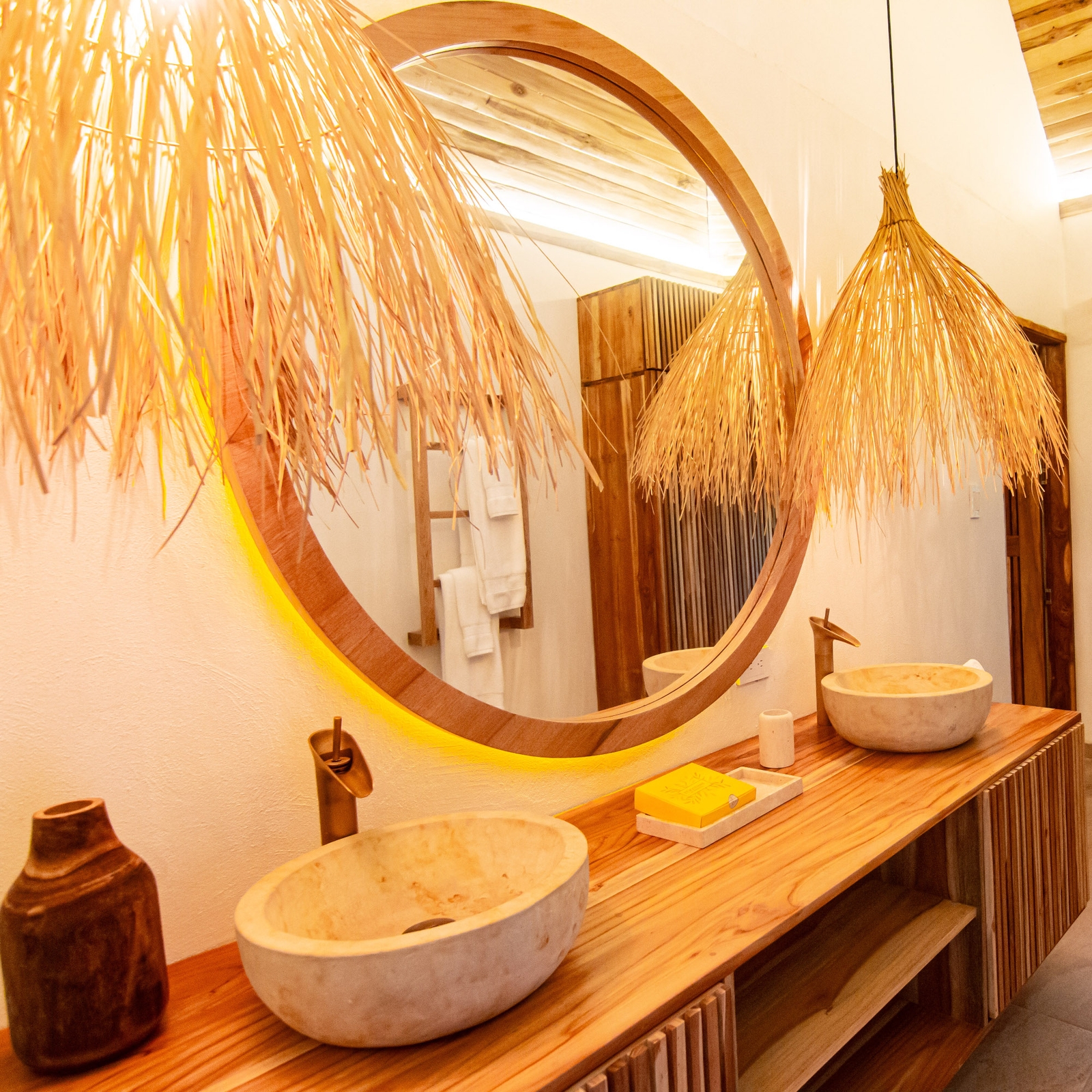 A bathroom vanity with a big round mirror, two marble sinks set on a wooden countertop with a dark bottle on the left. Overhead are two lamp shades made of brown grass.