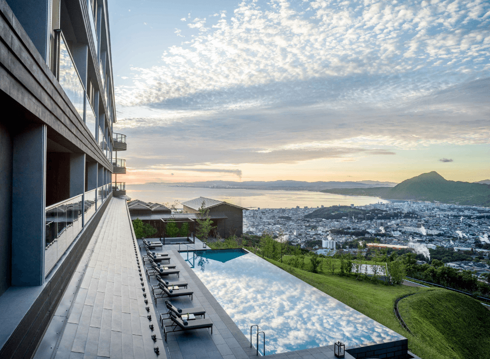 Wide view of a hotel and infinity pool overlooking mountains and the sea at sunset