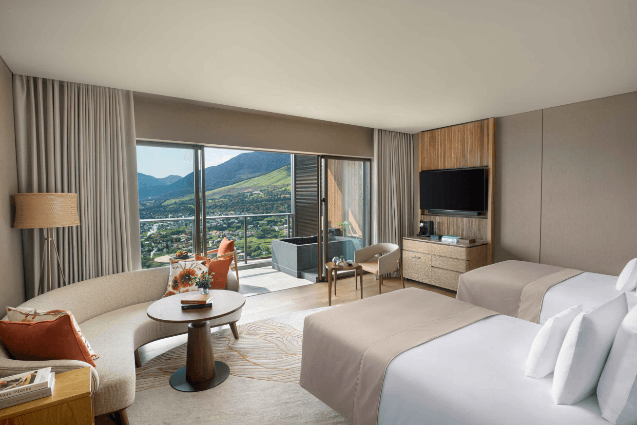 A bedroom with beige and white decor, two beds, a sofa and a view of the mountains
