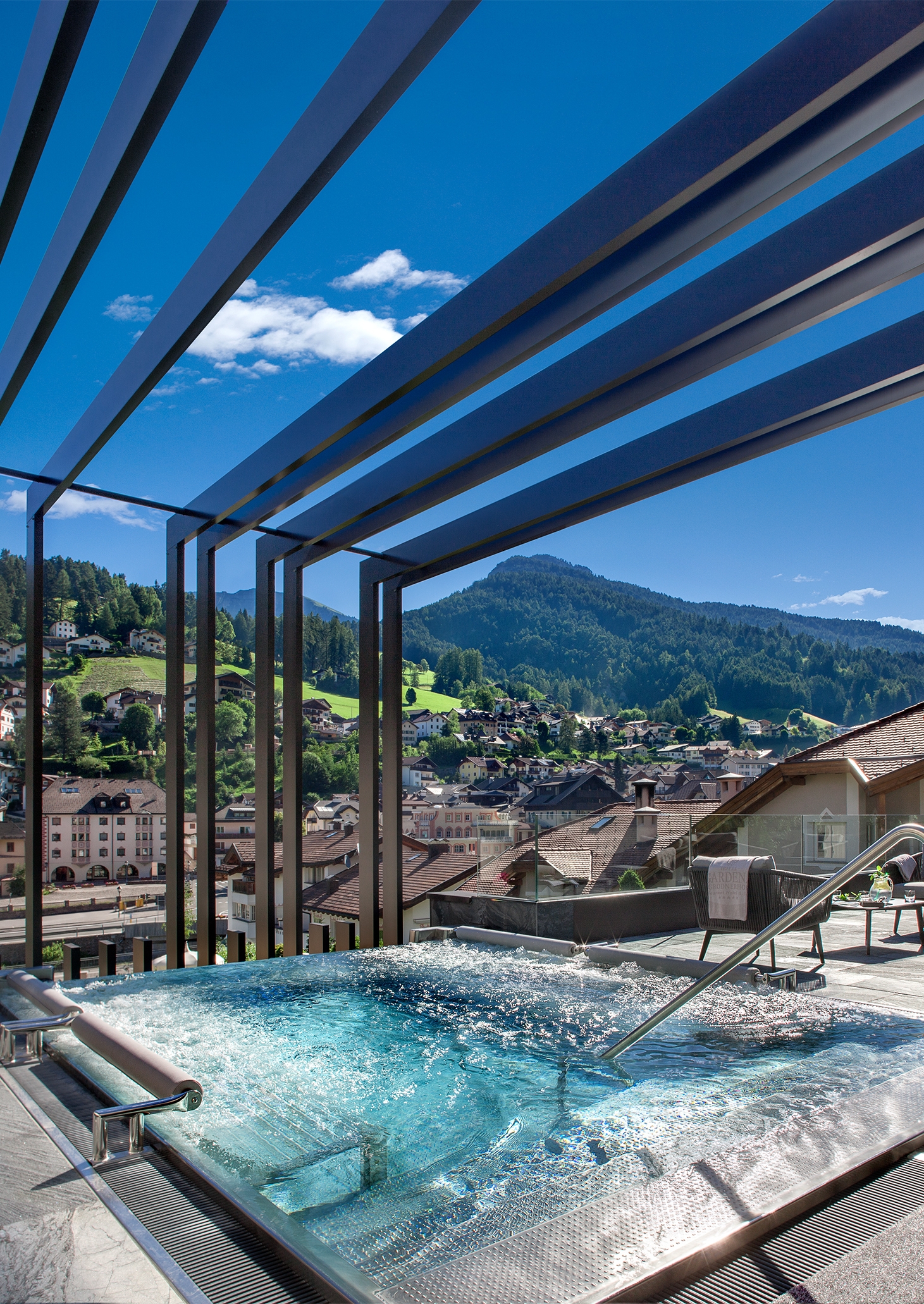 Hot tub with turquoise water and black shade panels above, and in the distance is the town with green mountainsides beyond and blue sky.