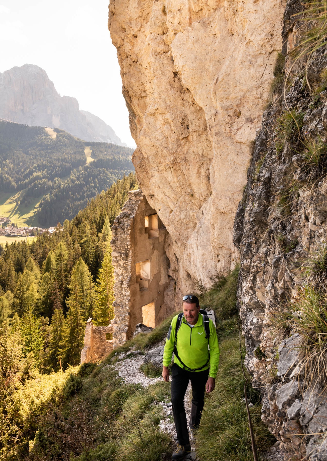 Man in a bright green shirt hiking up a narrow mountainside with tan rocks above and mountains in the distance behind him.