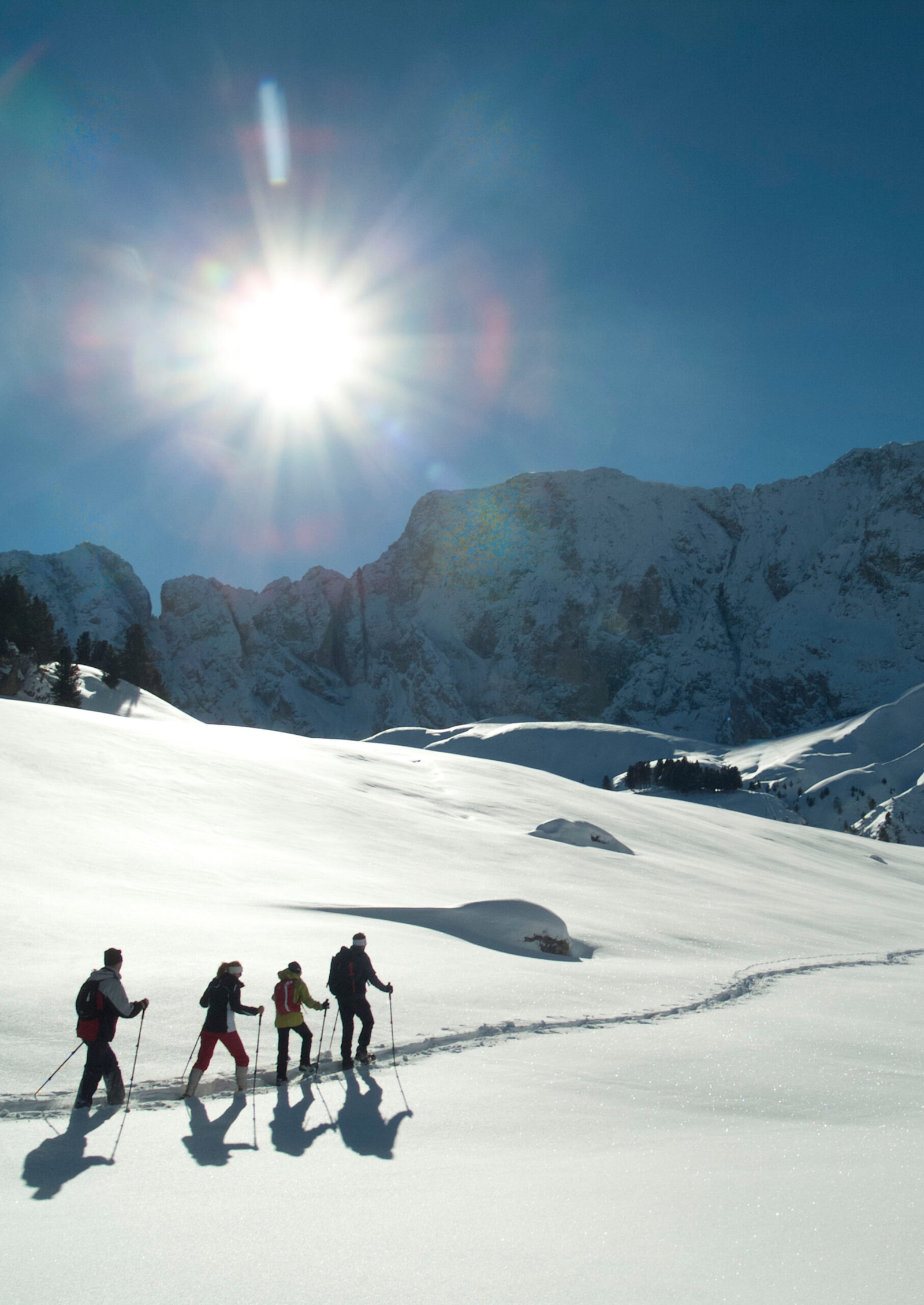 Shot of four people snowshoeing through a snowy mountain landscape with a bright sun shining over them.