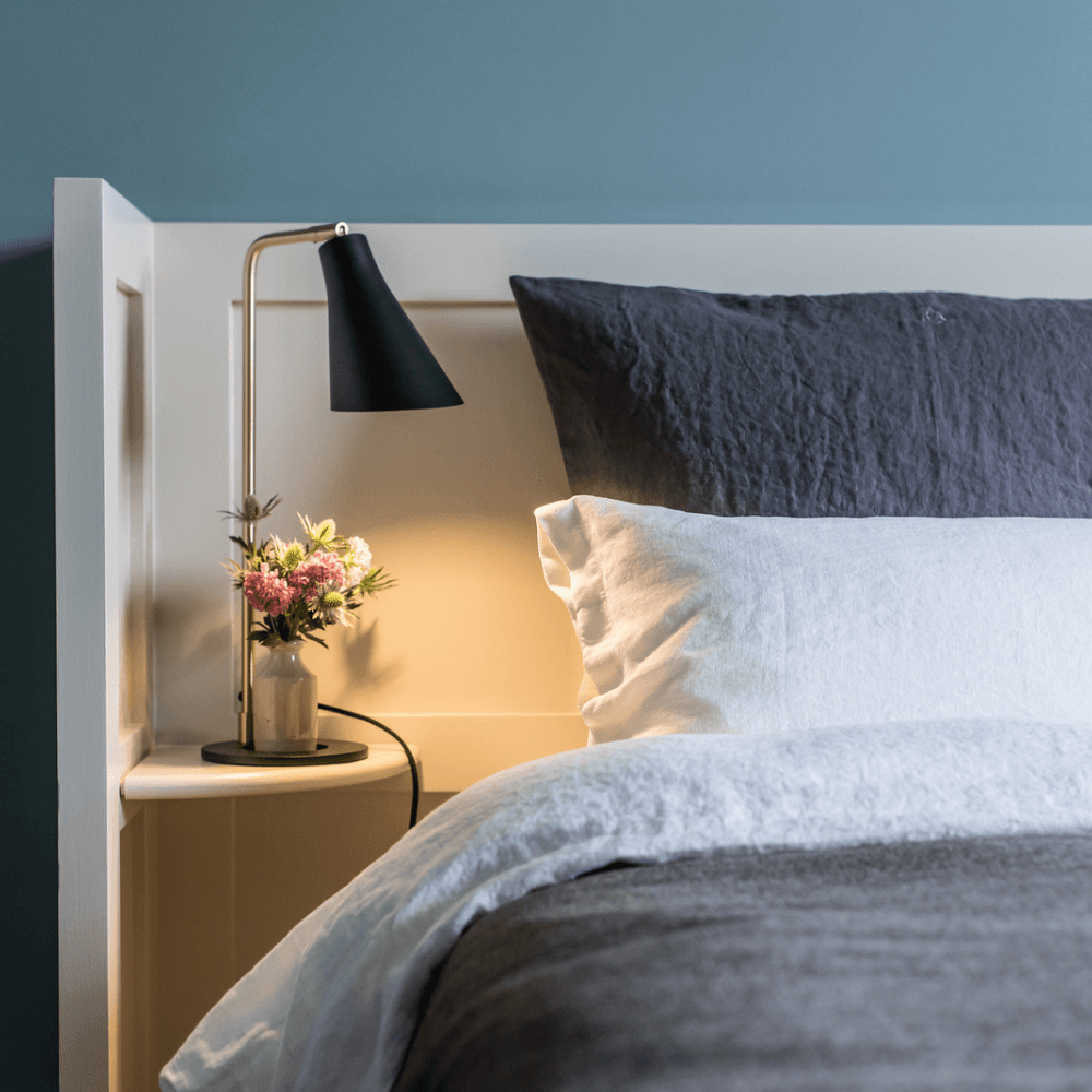 Close-up of a bedside shelf with a vase of flowers and a small lamp next to a dark grey bedspread and grey and white pillows against a white headboard