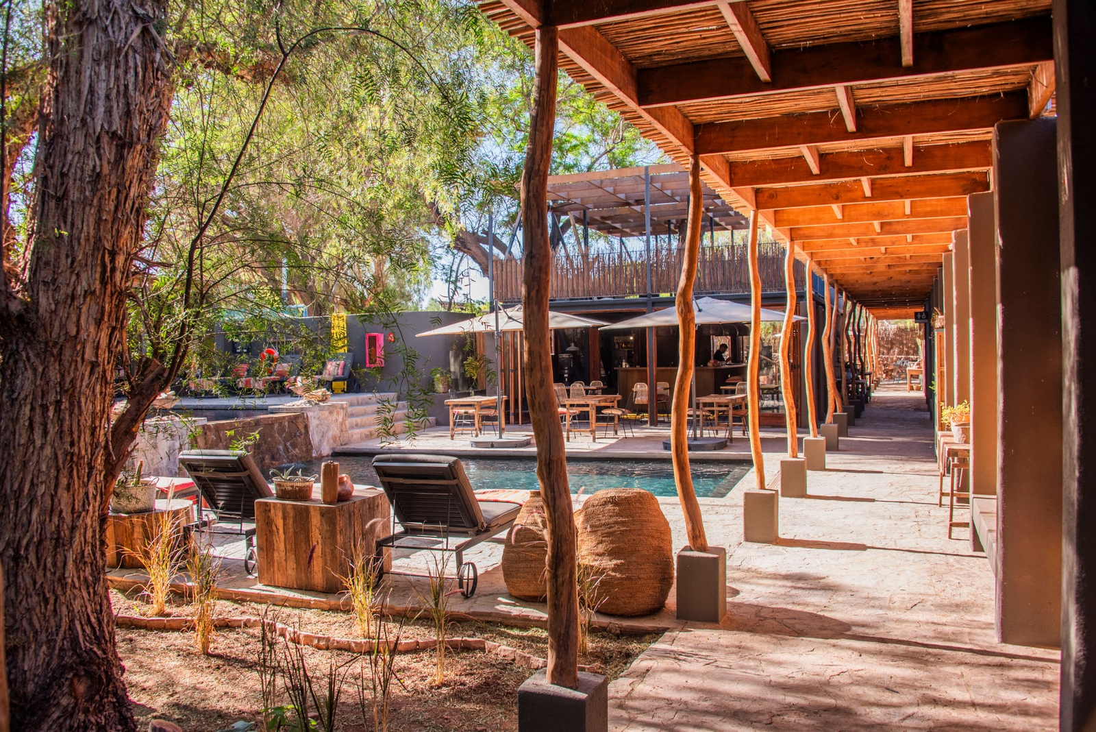 Exterior of a pool area with umbrella shades and an indoor-outdoor shaded walkway made of stone and covered by wooden portal, with tree branches for columns.
