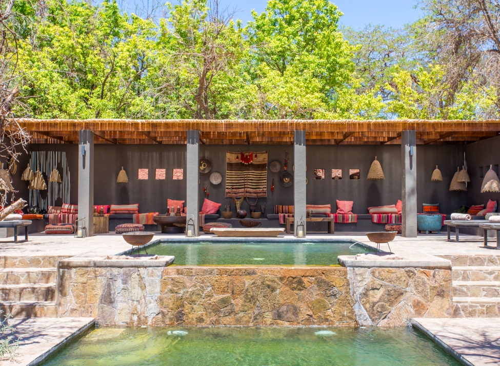 A patio terrace scene with two-tiered green pools and rock slab terraces with steps up to a shaded seating area with colourful sofas, chairs and lampshades in red, turquoise and brown, with green trees behind.