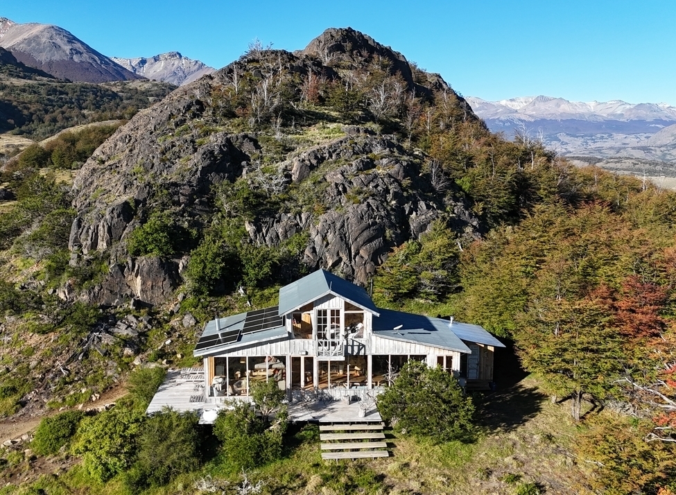 Two-storey cabin with pitched grey roof and steps leading up to the front sits in front of a large grey rock formation on a green mountain slope with mountains in the background.