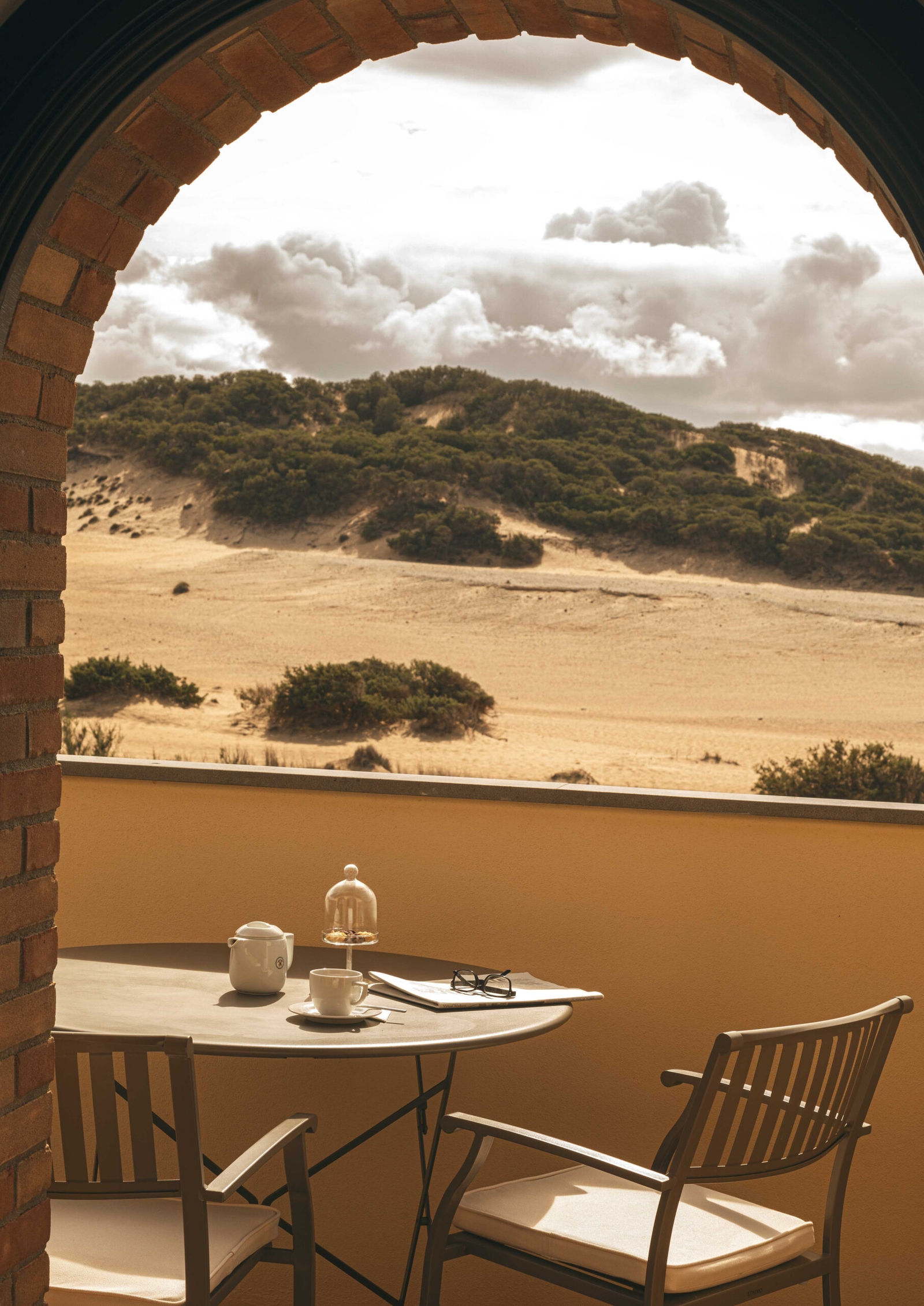 Exterior of a terrace with a table and chairs overlooking the dunes, shot through a stone archway.