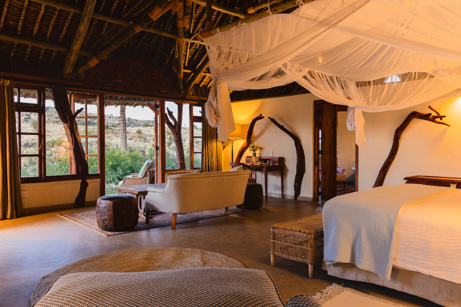 A safari bedroom with a bed with white mosquito netting, a sofa and wooden thatch ceiling and windows looking out onto the bush.