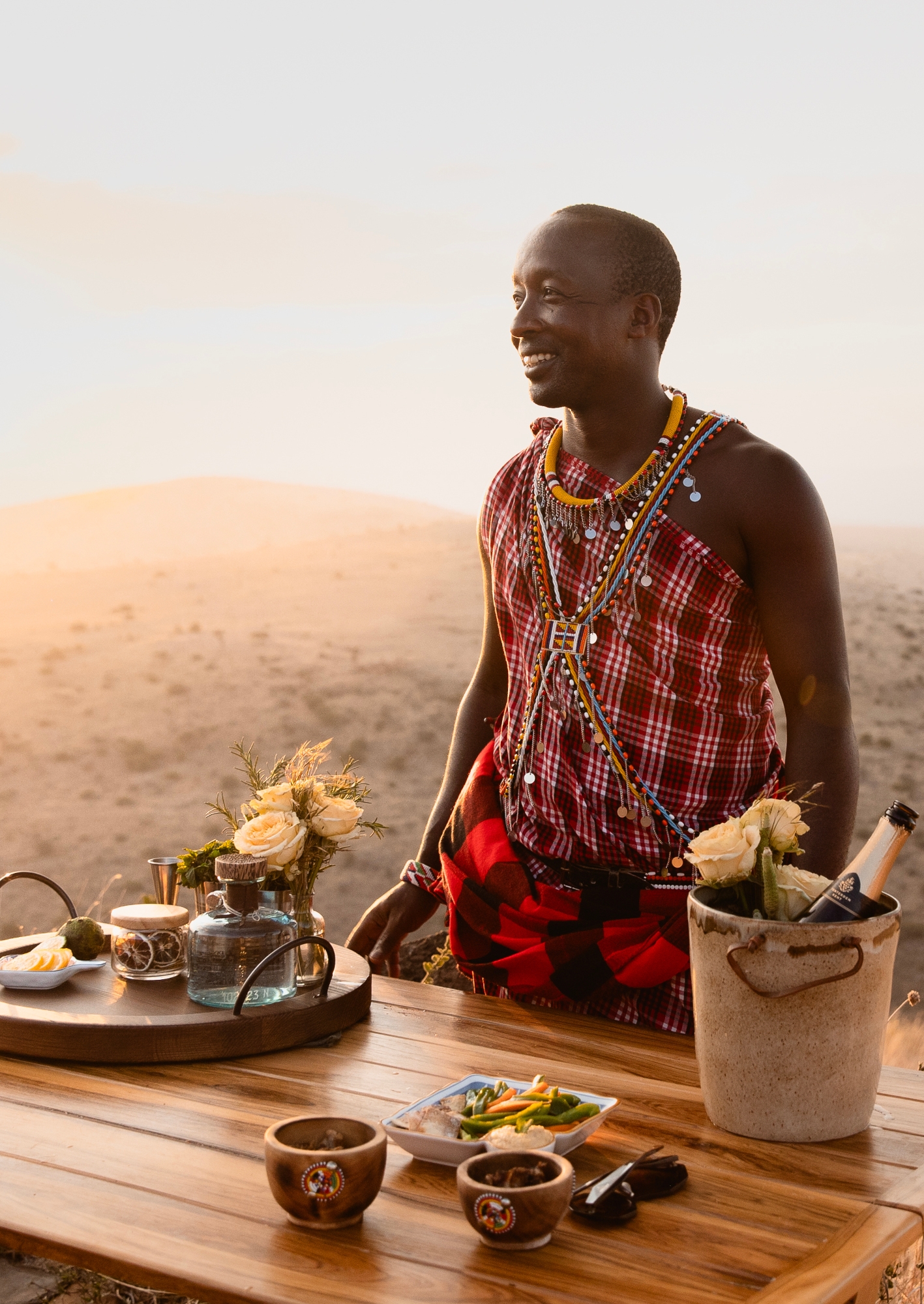 A man standing behind behind a table with drinks service and a sunset and African bushland in the background