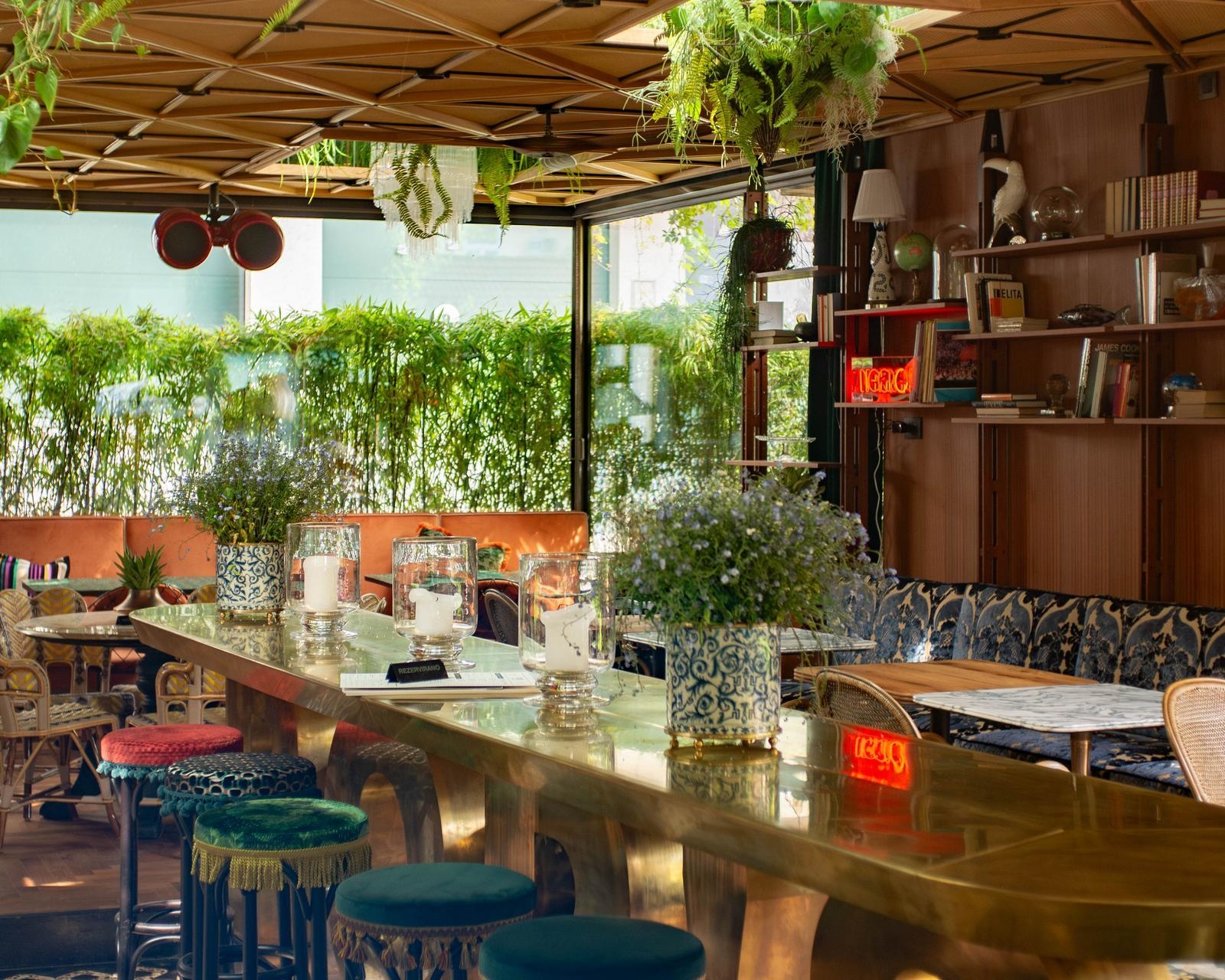Trendy bar interior with a long brass counter, patterned blue and green stools, and lush hanging plants.