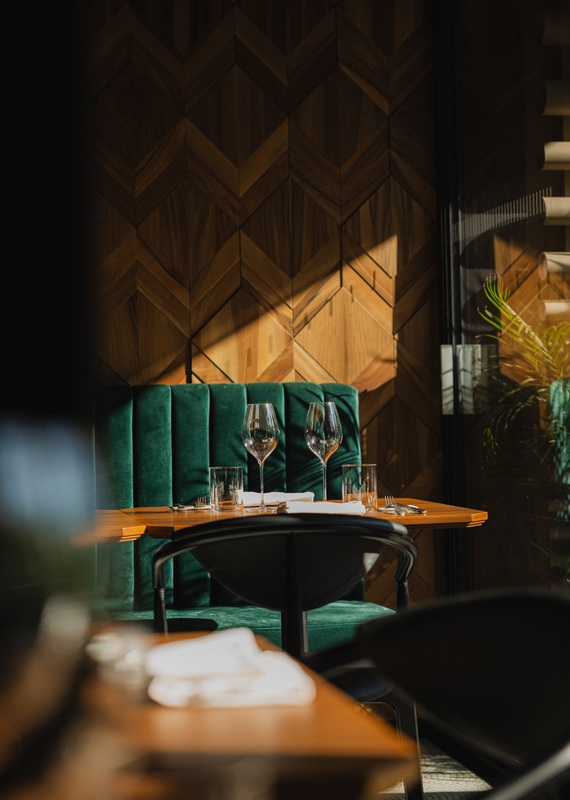 Close-up of a green velvet restaurant booth and wooden table with wine glasses against a geometric wood wall.