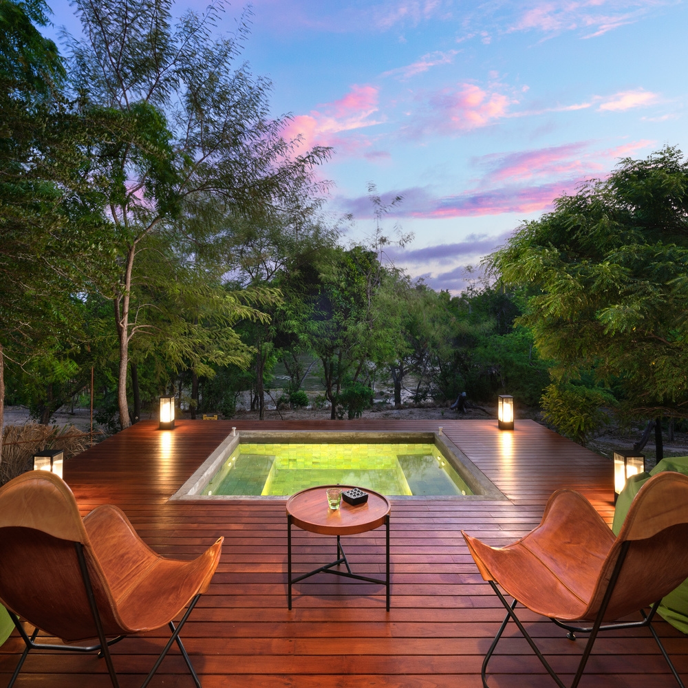 An illuminated square plunge pool set in a wooden deck with two leather chairs facing a lush forest at sunset.
