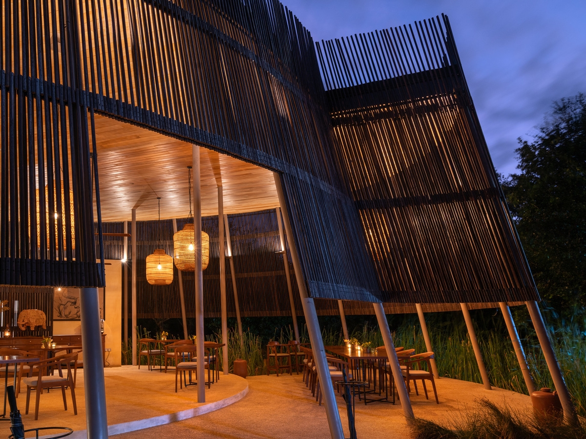 A modern open-air restaurant at night with slatted wood walls, warm glowing lanterns, and wooden dining tables.