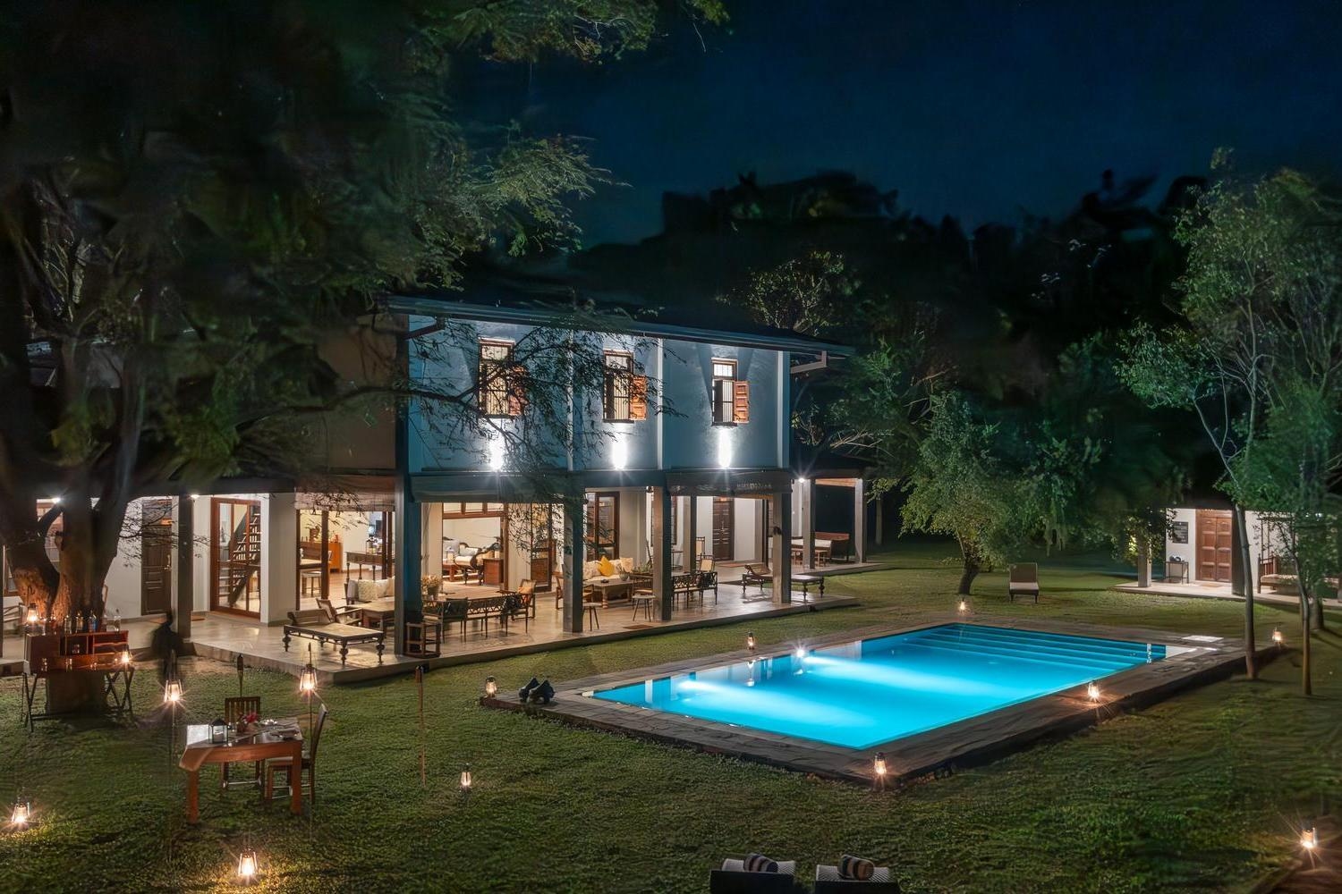 Large villa with a glowing blue swimming pool at night, surrounded by lush green lawn and trees.