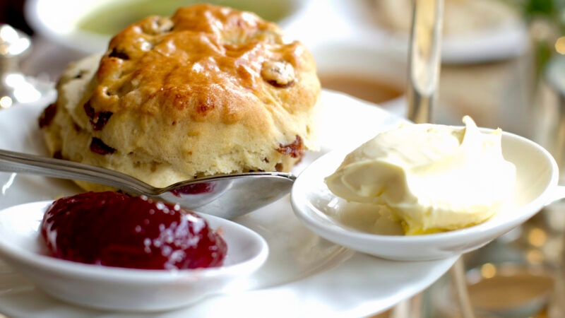 A golden-brown fruit scone served with a side of red strawberry jam and thick clotted cream on white chinaware.