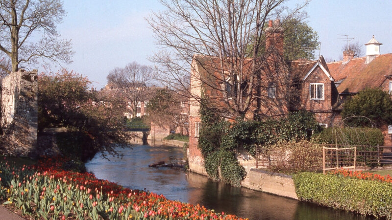 A brick house next to a flowing river with a large bed of blooming red and yellow tulips in the foreground.