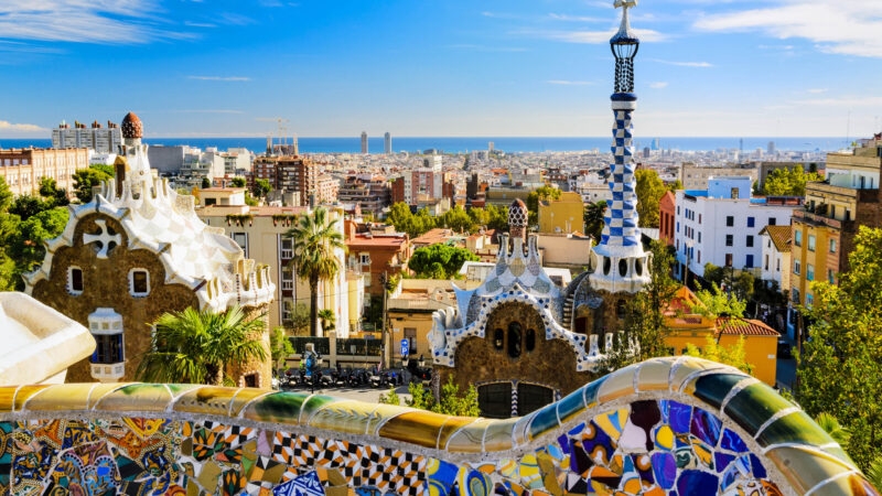 View of Park Guell's mosaic walls and iconic buildings with the Barcelona city skyline in the background.
