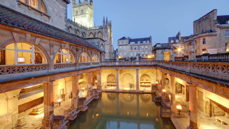 The Roman Baths in Bath, England, with glowing torches and stone pillars reflecting in the thermal water.