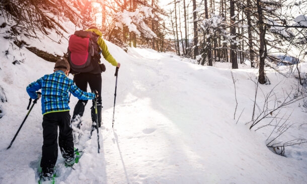Back view of an adult with a red backpack and a child in a blue plaid jacket snowshoeing up a snowy hill in a forest.