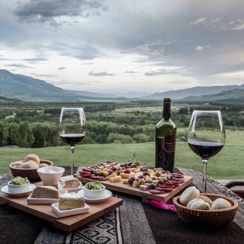 A table set with wine, glasses, cheese, meats, and bread, overlooking a wide green valley and mountains. Luxury Argentina trips.