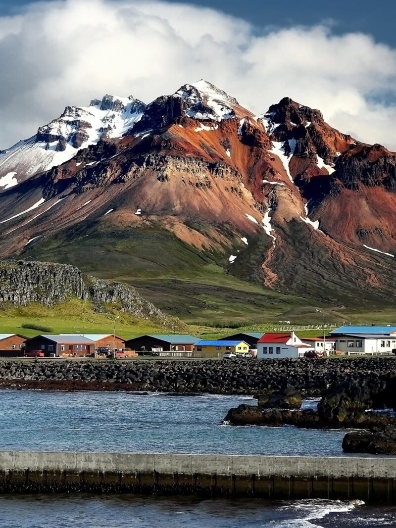 A row of colorful coastal houses sits on the rocky shore beneath a large, red, snow-capped mountain, ideal for luxury Iceland holidays.