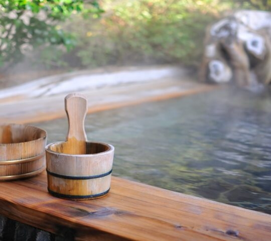 Wooden water buckets rest on the edge of a steaming outdoor onsen bath with a stone and foliage background.