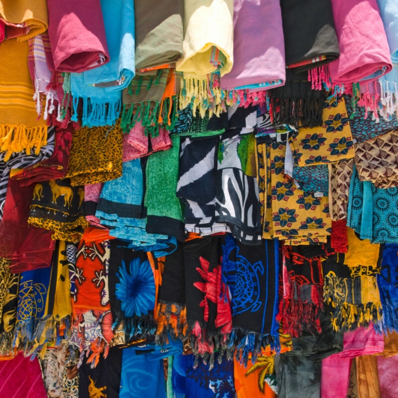 Luxury Kenya Safari Tours - Rows of brightly colored, patterned fabrics with fringes hanging vertically in a dense marketplace arrangement.