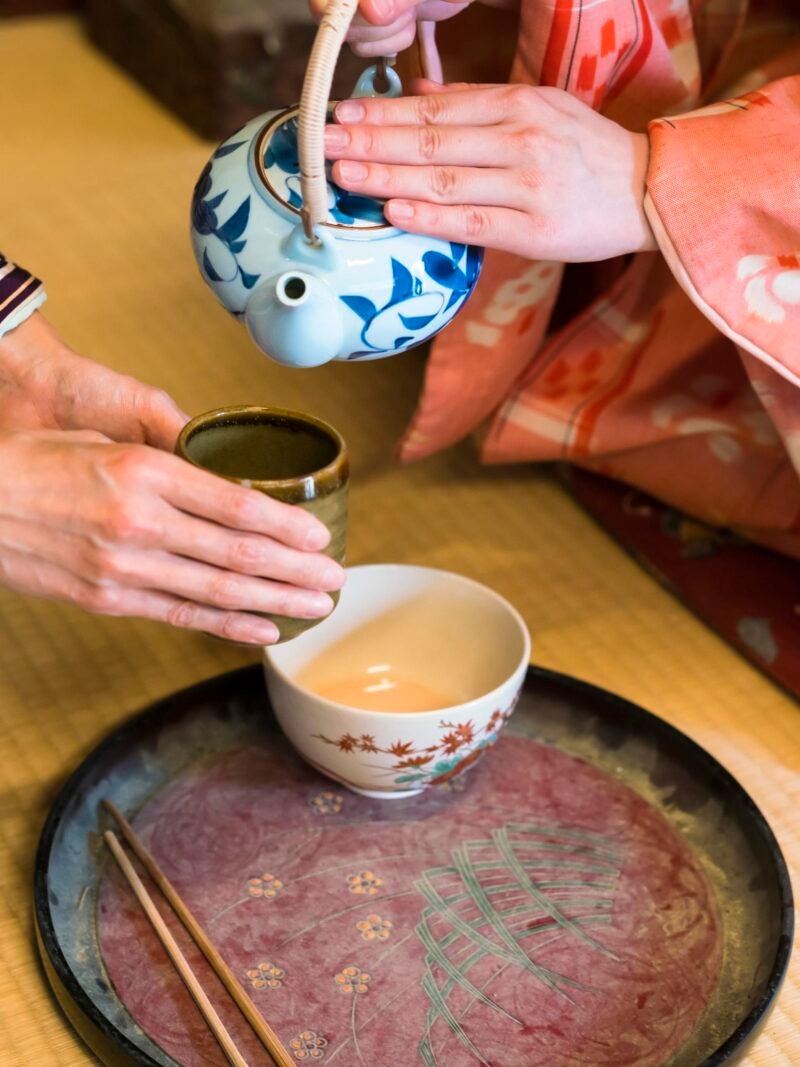Private Japan Luxury Tours - detail of two women in traditional kimono, kneeling on tatami preparing, pouring a cup of tea which is in the hands of one woman. They are in traditional Japanese old house on tatami.