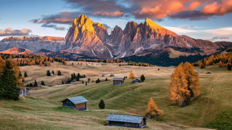 Rustic wooden huts in a mountain valley at sunset with glowing orange clouds and jagged peaks in the background.