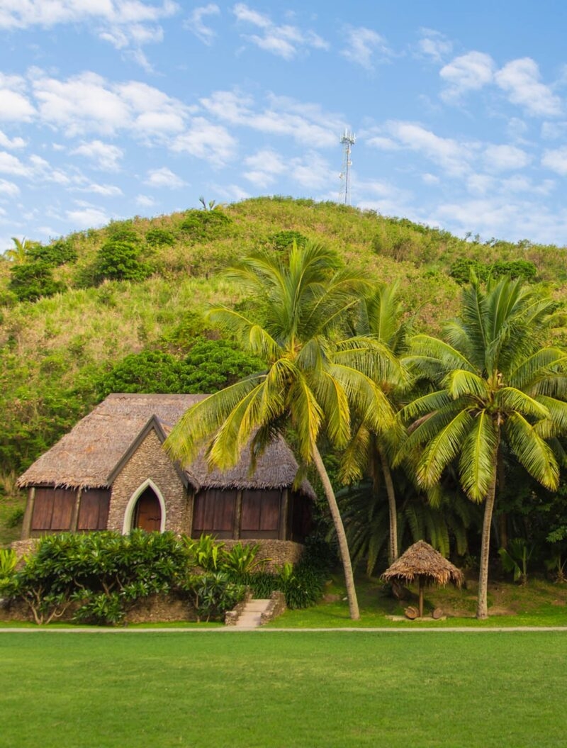 Traditional thatched-roof building and palm trees at the base of a lush green hill during luxury Fiji vacations.