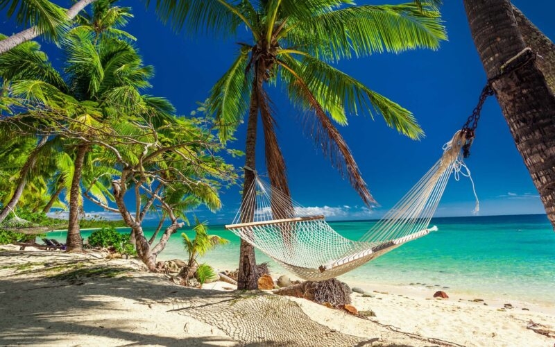 Rope hammock tied to palm trees on a white sand beach overlooking a tropical blue sea for luxury Fiji vacations.