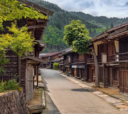 A quiet, historic street in Tsumago-juku, Japan, lined with traditional dark wood buildings and forested hills in the background.