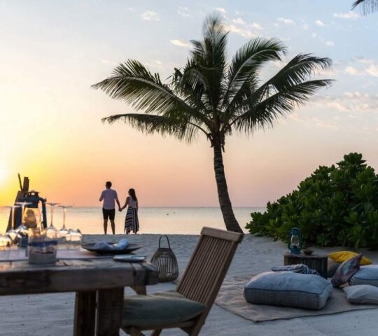 Silhouette of a couple holding hands on a tropical beach at sunset near a dinner table and floor cushions.
