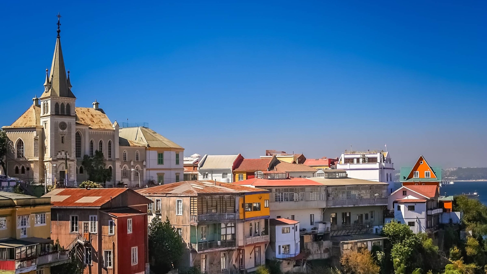 Colorful houses on a hillside next to a large stone church with a tall spire under a clear blue sky.