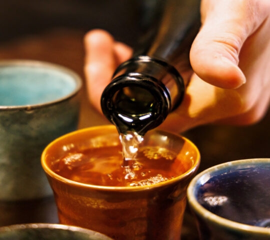 Close-up of sake being poured from a bottle into a small brown ceramic cup among other blue cups.