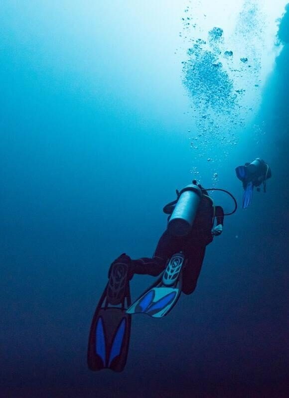 Two scuba divers with tanks and fins swim through deep blue water towards a source of light with rising bubbles.