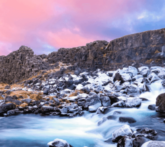 A serene river flowing over rocks with a pink and blue twilight sky in the background.