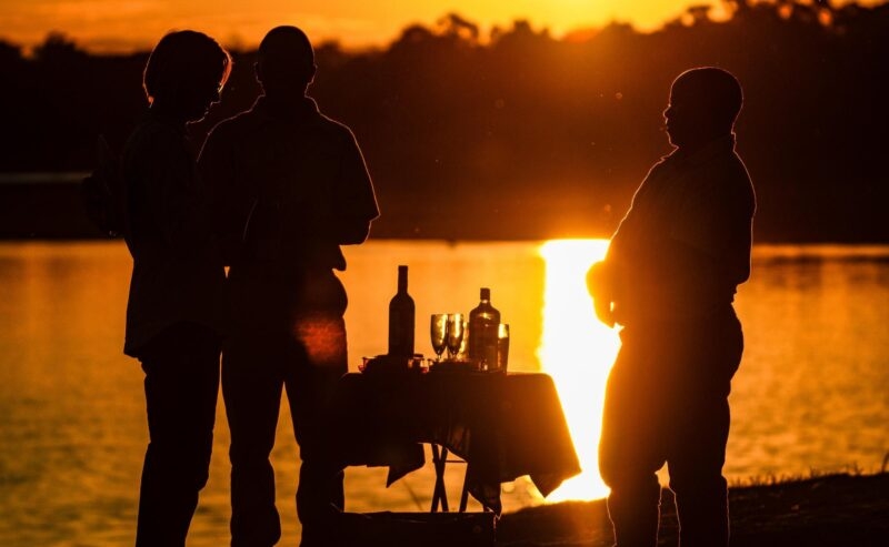 Silhouettes of three people standing by a table with bottles and glasses at sunset by a river.