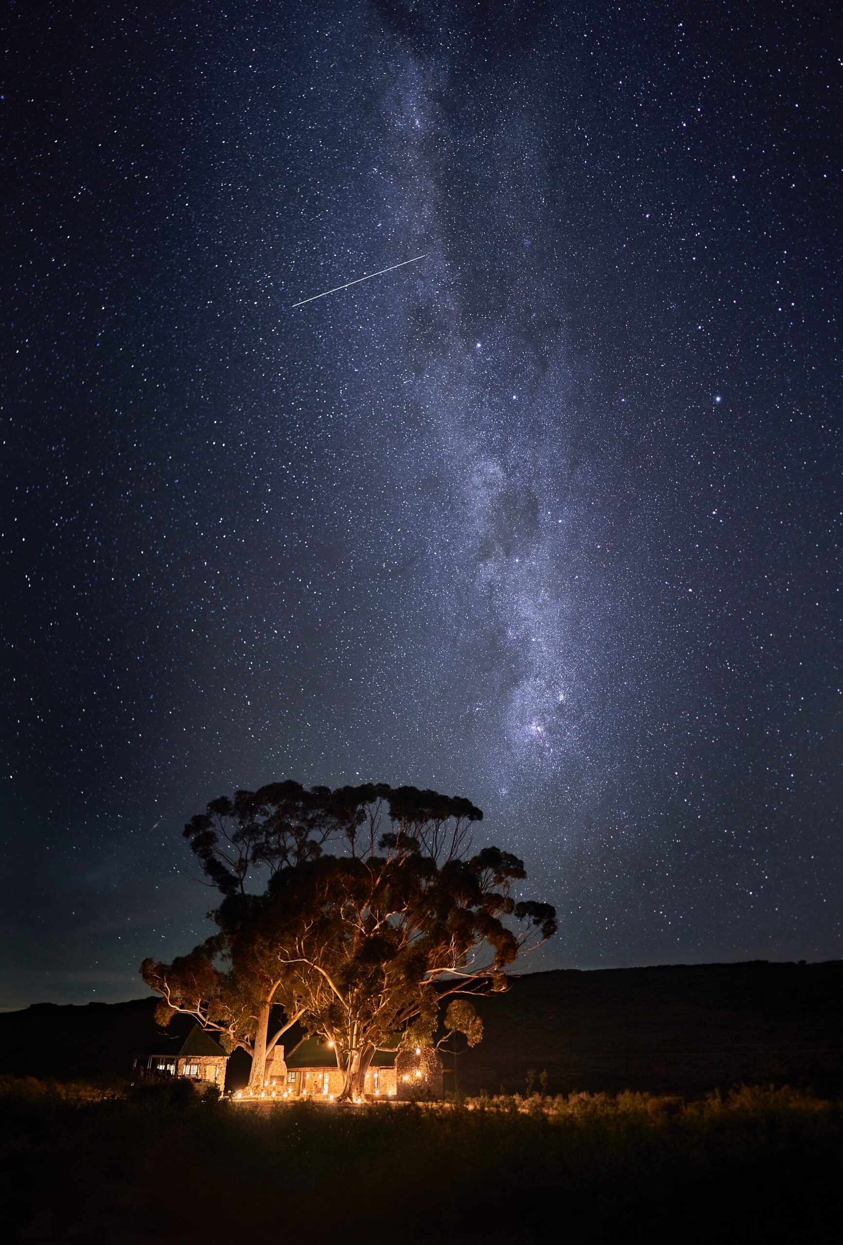 Illuminated house under a starry sky with the Milky Way and a shooting star.