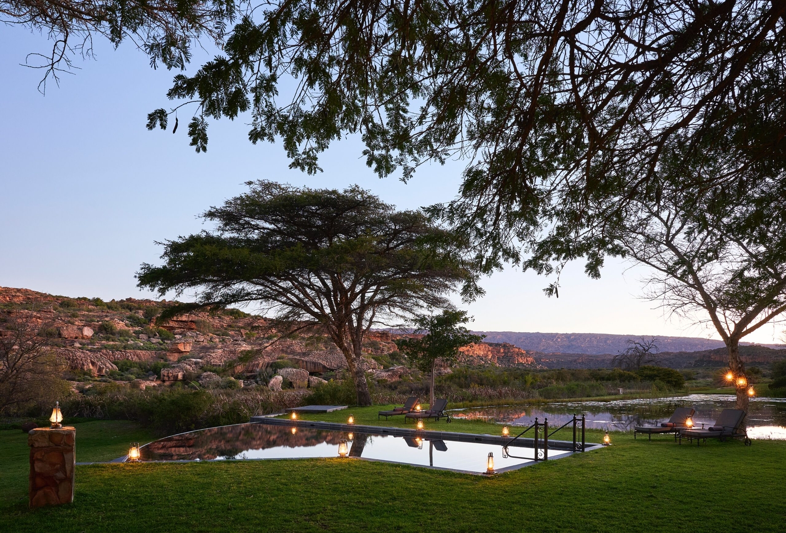 Tranquil outdoor pool with lanterns at dusk, surrounded by lush trees and rocky hills.