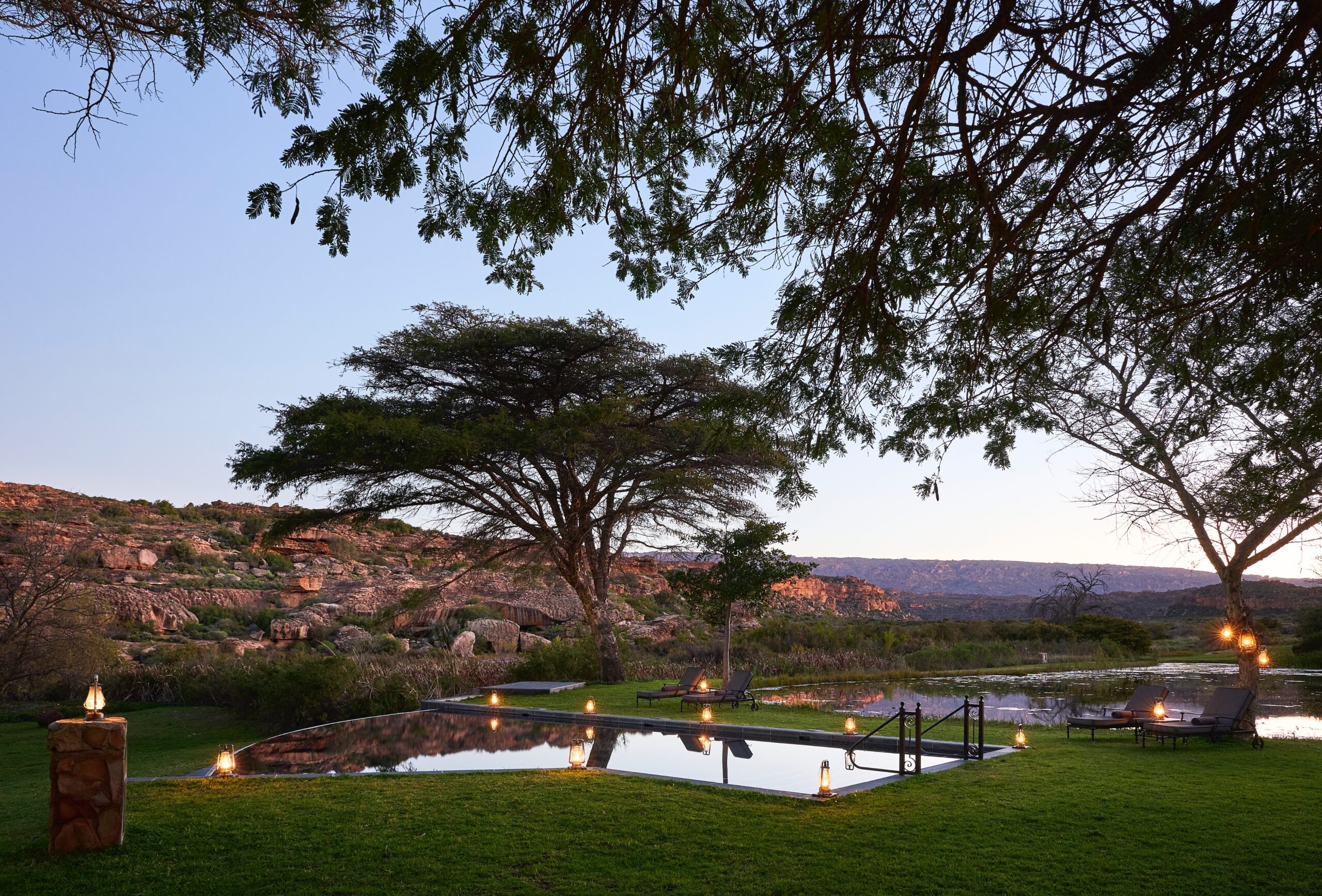 Tranquil outdoor pool with lanterns at dusk, surrounded by lush trees and rocky hills.