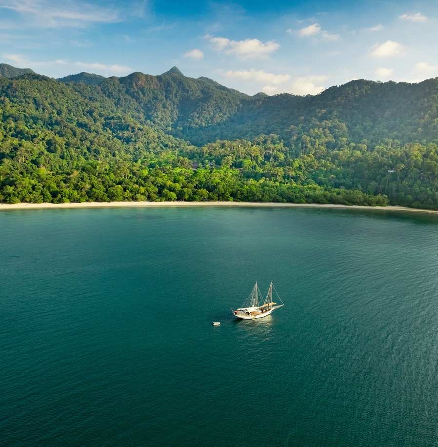 Aerial view of a sailboat anchored in a clear blue bay surrounded by high green mountains.